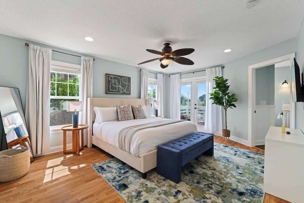 A bedroom with a bed, a ceiling fan, a window with curtains, a bedside table, and a plant.