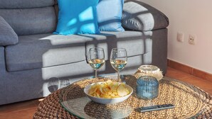 A living room with a grey sofa, two blue cushions, a glass coffee table with snacks, two wine glasses, and a remote control.