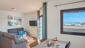 A modern living room with a grey sofa, a dining table with breakfast items, and a large window offering a view of a beach and mountains.