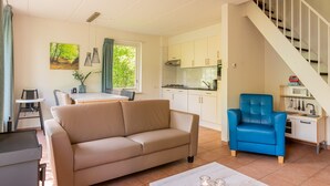 Un salon avec un canapé beige et un fauteuil bleu, un coin repas avec une table et des chaises, une cuisine avec des meubles blancs et une cuisinière, et un escalier avec des rampes blanches.