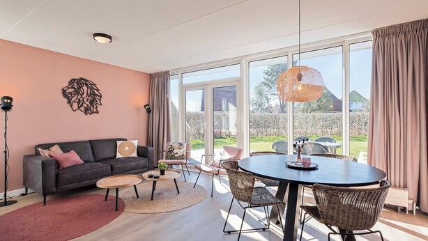 A modern living room with a grey sofa, round coffee table, wicker chairs, and a large window offering a view of the outdoors. - Semi-detached bungalow for 5 persons (Vlagtwedde)