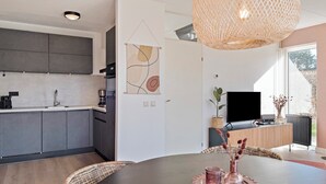A modern kitchen with dark cabinets, a white countertop, and a coffee maker. There is a dining area with a round table, wicker chairs, and a vase with dried flowers. A television is mounted on the wall, and a woven light fixture hangs above the dining table. - Semi-detached bungalow for 5 persons (Vlagtwedde)