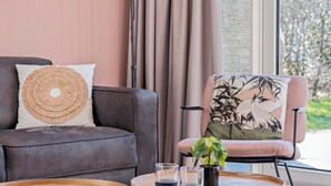 A modern living room with a grey sofa, a round woven pillow, a chair with a bird print cushion, and a small table with glasses and a plant. - Semi-detached bungalow for 5 persons (Vlagtwedde)