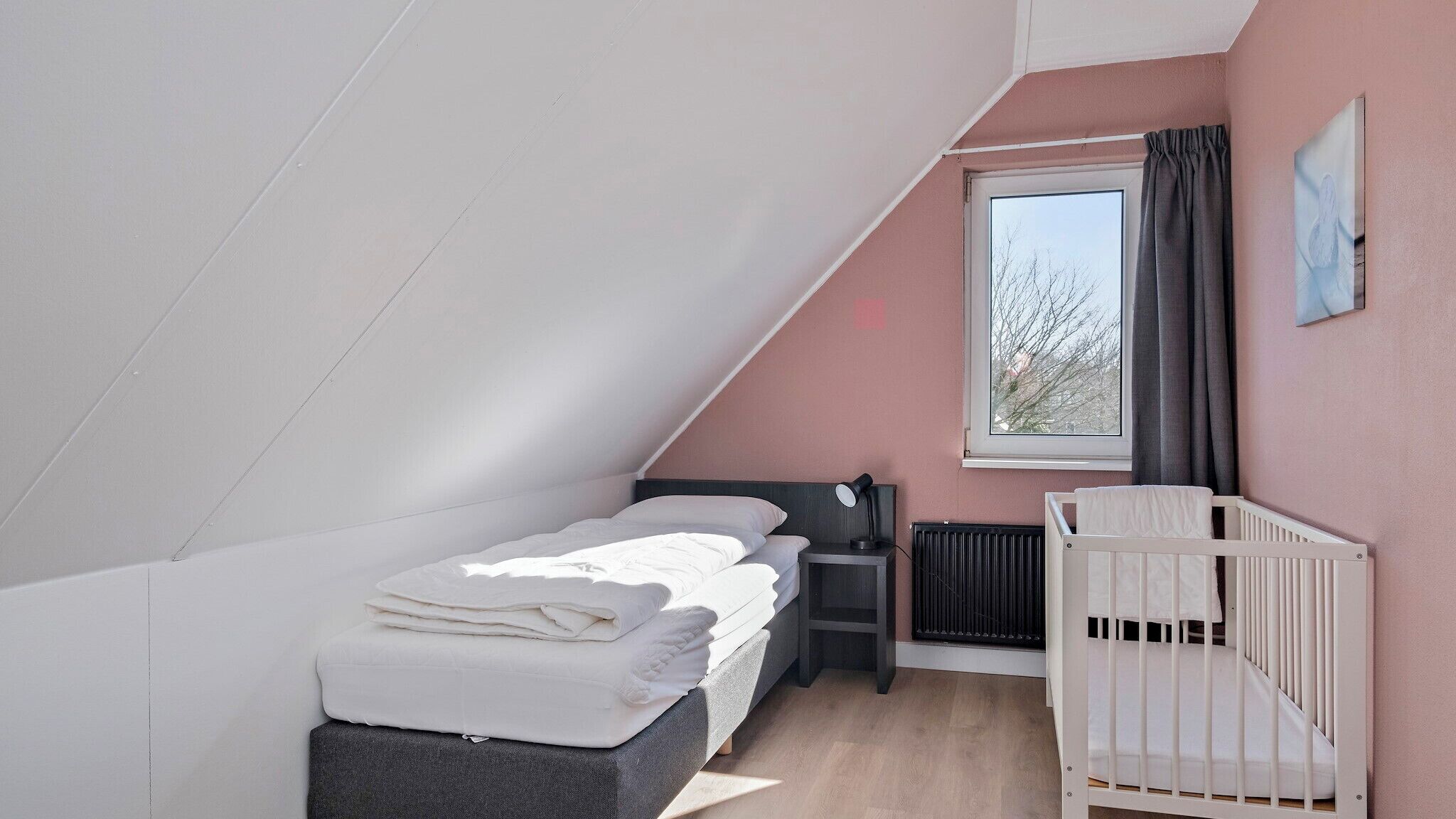 A bedroom with a sloped ceiling, a single bed, a nightstand, a crib, and a window with curtains.