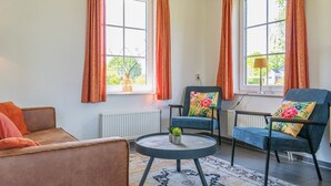 A living room with two armchairs, a sofa, a round coffee table, and a view of the outdoors through large windows with orange curtains. - Detached 6-person bungalow (Vlagtwedde)