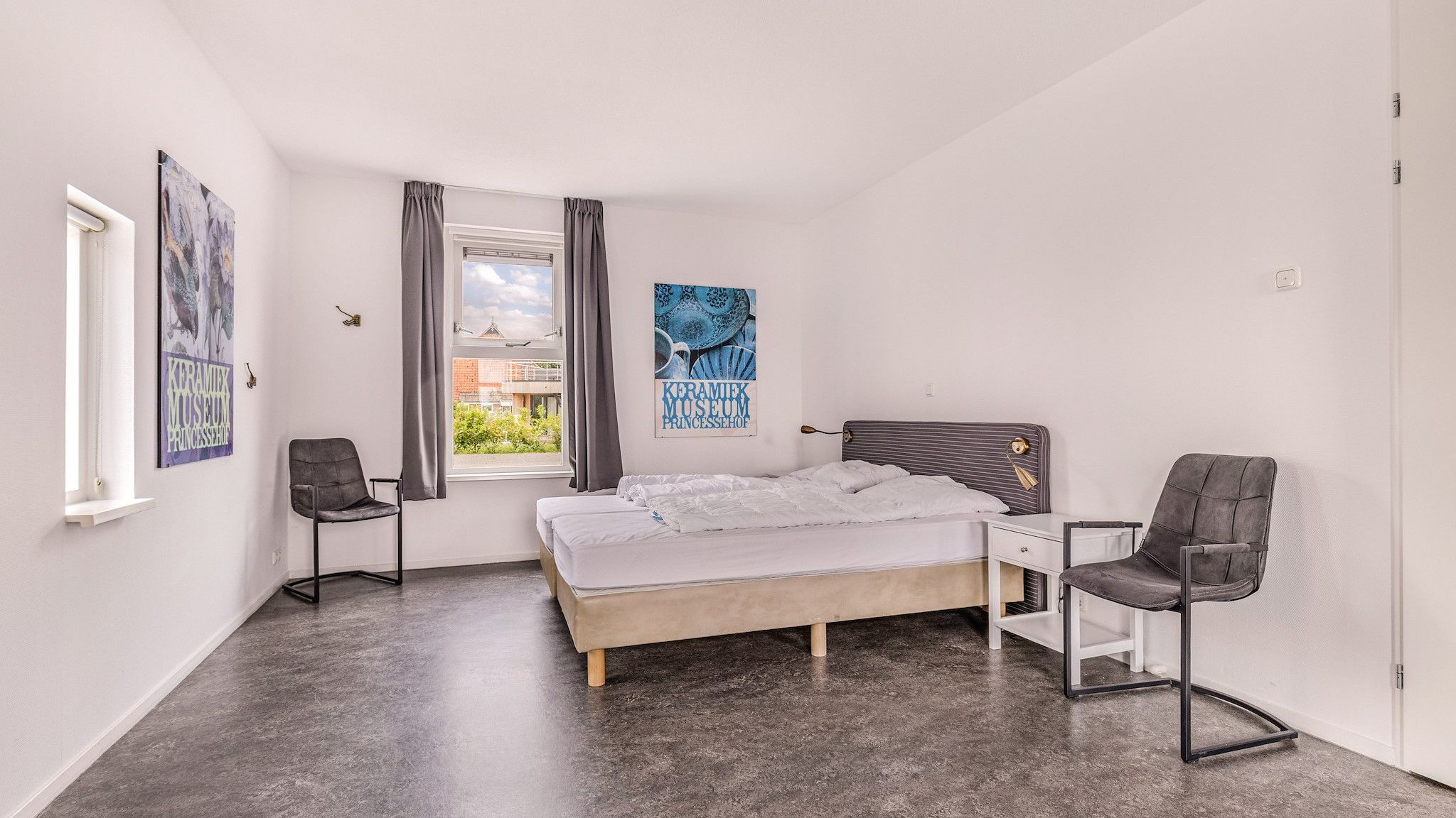 Une chambre à coucher avec un lit, deux chaises, une fenêtre avec des rideaux et deux affiches au mur.
