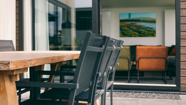 A wooden dining table with a black mesh chair in front of a glass door leading to a patio area with a painting and a sofa.