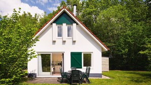 Ein zweigeschossiges Haus mit einem grünen Dach, weißen Außenwänden und einer Schiebetür, die auf eine Terrasse mit Tisch und Stühlen führt.