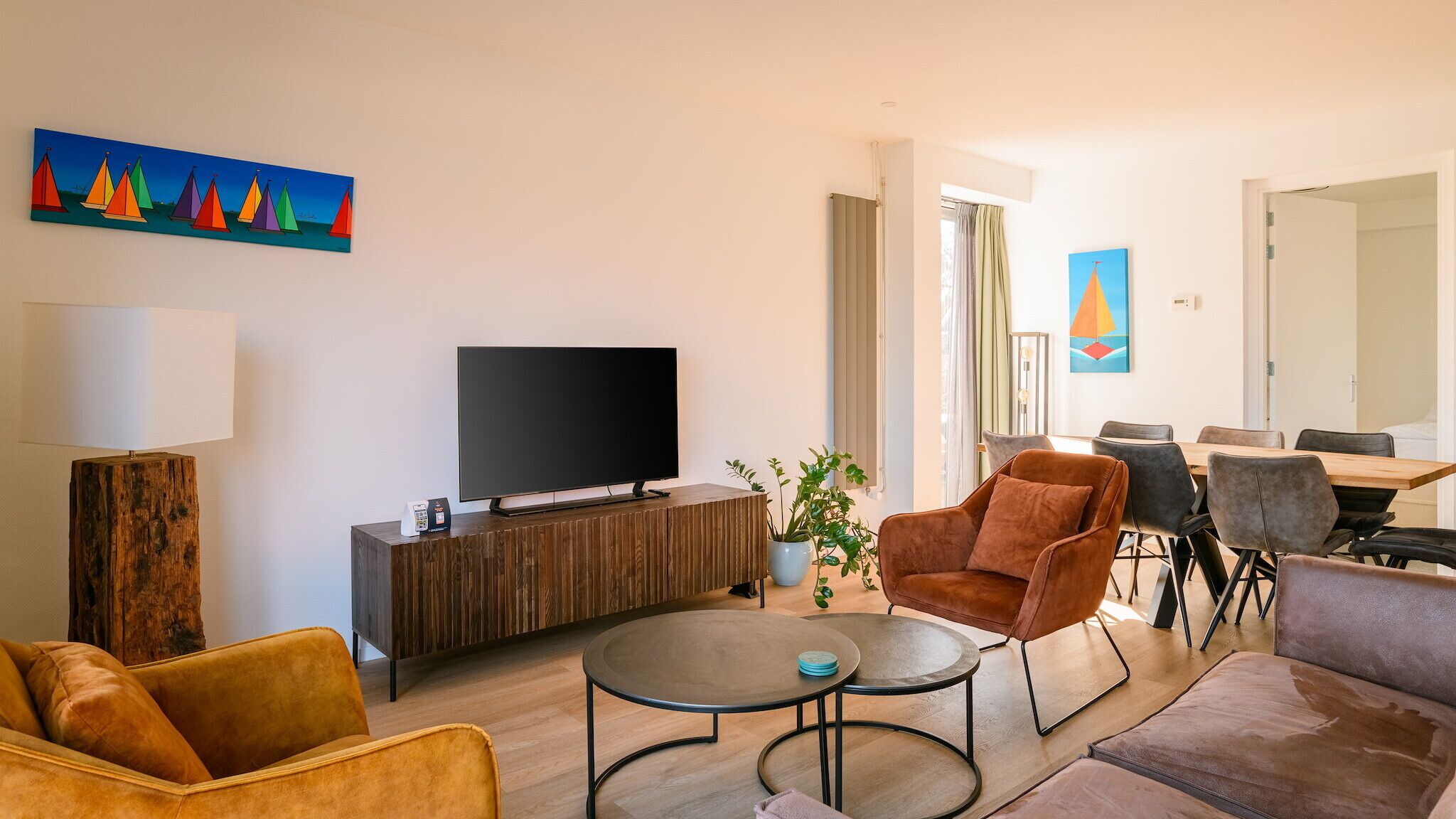 A modern living room with a flat-screen TV, a wooden cabinet, a coffee machine, a sofa, a dining table with chairs, and a painting on the wall.
