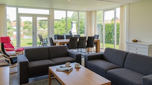 A spacious living area with a large window overlooking a garden and a lake. There are two sofas, a dining table with chairs, and a red chair. - Detached bungalow for 8 persons (Vlagtwedde)