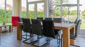 A dining area with a wooden table and black chairs, large windows with a view of greenery and buildings, and a red chair. - Detached bungalow for 8 persons (Vlagtwedde)