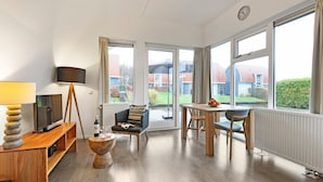 Un salon moderne avec un téléviseur, des meubles en bois, une lampe et de grandes fenêtres donnant sur l’extérieur.