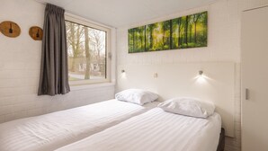 Une chambre avec deux lits simples, une grande fenêtre avec des rideaux, un tableau représentant une forêt et un mur en briques.