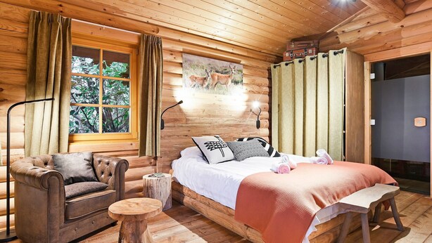 Ein gemütliches Schlafzimmer in einer Holzhütte mit Bett, Stuhl, kleinem Tisch und einem Fenster mit Vorhängen.