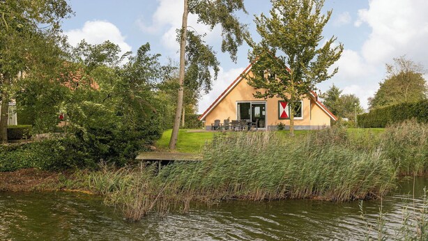 A house with a red roof and a flag on the door, surrounded by greenery and a body of water. - Detached bungalow for 6 persons (Tynaarloo)