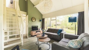 A spacious living room with a sofa, coffee table, and TV, a staircase, and large windows with curtains. - Semi-detached 6-person bungalow (Midlaren)