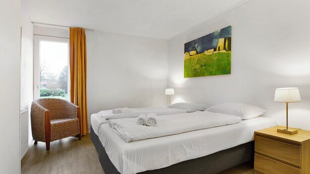 A hotel room with a bed, two bedside lamps, a chair, a painting on the wall, and a window with curtains. - Semi-detached 6-person bungalow (Midlaren)