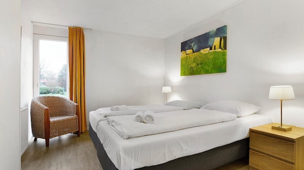 A hotel room with a bed, two bedside lamps, a chair, a painting on the wall, and a window with curtains.