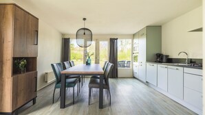 A modern kitchen with white cabinets, a dining table with chairs, and a view through the window. - Semi-detached 6-person bungalow (Midlaren)