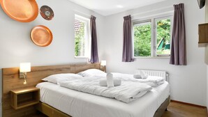 Een hotelkamer met een bed, twee nachtkastjes, een raam met gordijnen en uitzicht op groen.