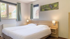A neatly made bed in a room with two windows, a radiator, and a wall-mounted picture of a bird. - Comfortable bungalow for 6 people (Diessen-Baarschot)