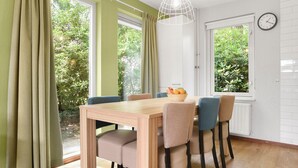 A dining area with a wooden table, six chairs, a fruit basket, and two windows with curtains. - Comfortable bungalow for 6 people (Diessen-Baarschot)