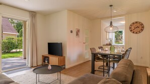 A modern living room with a flat-screen TV, a dining table with chairs, a sofa, and a large wall clock. - Comfortable bungalow for 4 persons (Diessen)