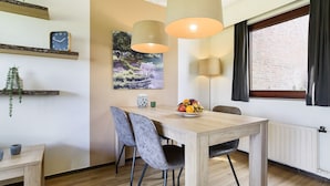 Uma sala de jantar com mesa de madeira, duas cadeiras, um cesto de frutas, um relógio de parede, um quadro e luminárias pendentes.