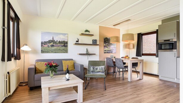 Sala de estar com sofá, mesa de centro com garrafa e flores, área de jantar com cadeiras e mesa, e cozinha com geladeira e forno.