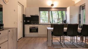 A modern kitchen with a dining area, featuring a built-in oven, a stove, and a window with a view of greenery. - Detached bungalow for 6 persons (Kootwijk)