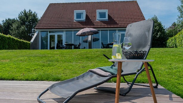 A reclining chair on a wooden deck with a table beside it, a bottle of water, a glass, and a basket. In the background, there is a house with a red-tiled roof and a glass door. - Bungalow Wellness 14 (Vlagtwedde)