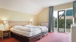 Une chambre d’hôtel avec deux lits, une tête de lit en bois, une table de chevet, une lampe et une vue sur un immeuble et de la verdure, le tout visible à travers une grande fenêtre.