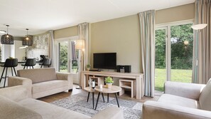 A modern living room with a sofa, coffee table, and TV. There is a dining area with chairs and a table, and large windows with curtains.