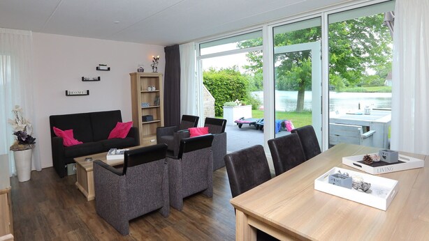 A modern living room with a black sofa, grey armchairs, a wooden coffee table, and a dining table with a white tray. A bookshelf and a window with a view of a lake are also visible. - Comfortable detached or semi-detached bungalow for 6 persons (Vlagtwedde)