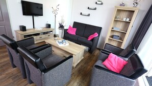 A modern living room with a black sofa, grey armchairs, a wooden coffee table, and a TV mounted on the wall. - Comfortable detached or semi-detached bungalow for 6 persons (Vlagtwedde)