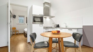 Eine kleine Küche mit Mikrowelle, Herd und modernen Geräten. Ein Esstisch mit Stühlen und eine Schale mit Obst sind vorhanden. Die Wände sind weiß und der Boden aus Holz.