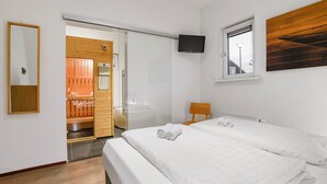 Ein Hotelzimmer mit Bett, Fernseher, Fenster und einem durchgegangene Tür erkennbaren Saunabereich.