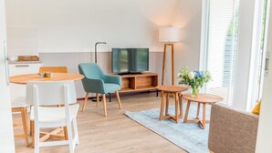 Ein modernes Wohnzimmer mit einem Holzesstisch, einem blauen Sessel, einem weißen Stuhl, einem Fernseher, einer Stehlampe und einem kleinen Beistelltisch mit Blumen in einer Vase.