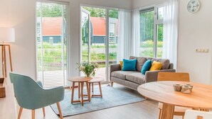 Ein modernes Wohnzimmer mit einer Couch, einem Esstisch und einem Stuhl. Große Fenster mit Jalousien, eine Uhr an der Wand und ein kleiner Tisch mit einem Blumenstrauß.