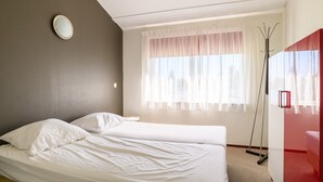 Ein Zimmer mit zwei Betten, einem Fenster mit durchsichtigen Vorhängen, einem runden Spiegel an der Wand und einer roten Akzentwand.