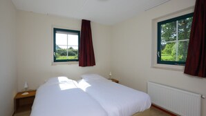 Ein Schlafzimmer mit einem Einzelbett, zwei Fenstern mit Vorhängen, einem Nachttisch mit Lampe und einer Heizung.