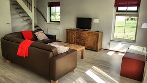 Ein Wohnzimmer mit einer Couch, einem Holzschrank, einem Fernseher und einem Fenster mit Blick ins Grüne.