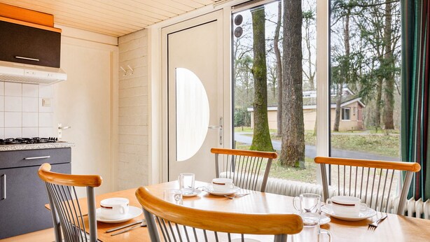 Un comedor con una mesa lista para dos personas, sillas, una ventana con vistas a árboles y casas, y una zona de cocina con armarios y una estufa.
