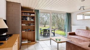 Sala de estar con estantería, televisor, sofá, mesa y puerta corrediza de cristal que da a un espacio exterior con sillas.
