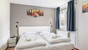 Un dormitorio con cama, dos mesitas de noche, una mesita auxiliar, una ventana con cortinas y un cuadro en la pared.