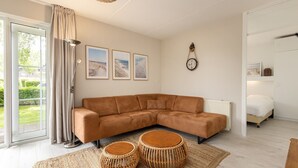 A modern living room with a sectional sofa, a large window with curtains, framed beach pictures, a wall clock, and a bed in the adjacent room.