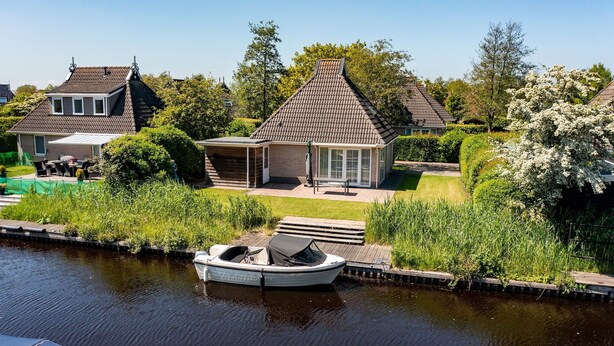 A canal-side house with a boat docked in the water, a patio with outdoor furniture, and a garden with various plants and trees.