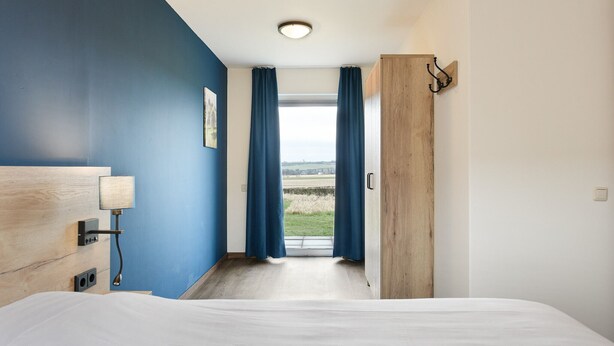 Quarto de hotel com cama, abajur, cabeceira de madeira, vista do campo através de uma janela com cortinas azuis, e porta de madeira com puxador.