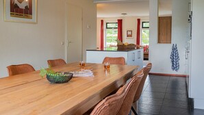 A dining area with a wooden table, chairs, and a bowl of fruit. There is a kitchen area in the background with a window and red curtains. - Detached bungalow for 9 persons (Vlagtwedde)