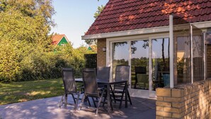An outdoor patio with a table and chairs, a brick wall, and a glass door leading to a covered area. - Comfortable bungalow for 4 persons (Diessen-Baarschot)